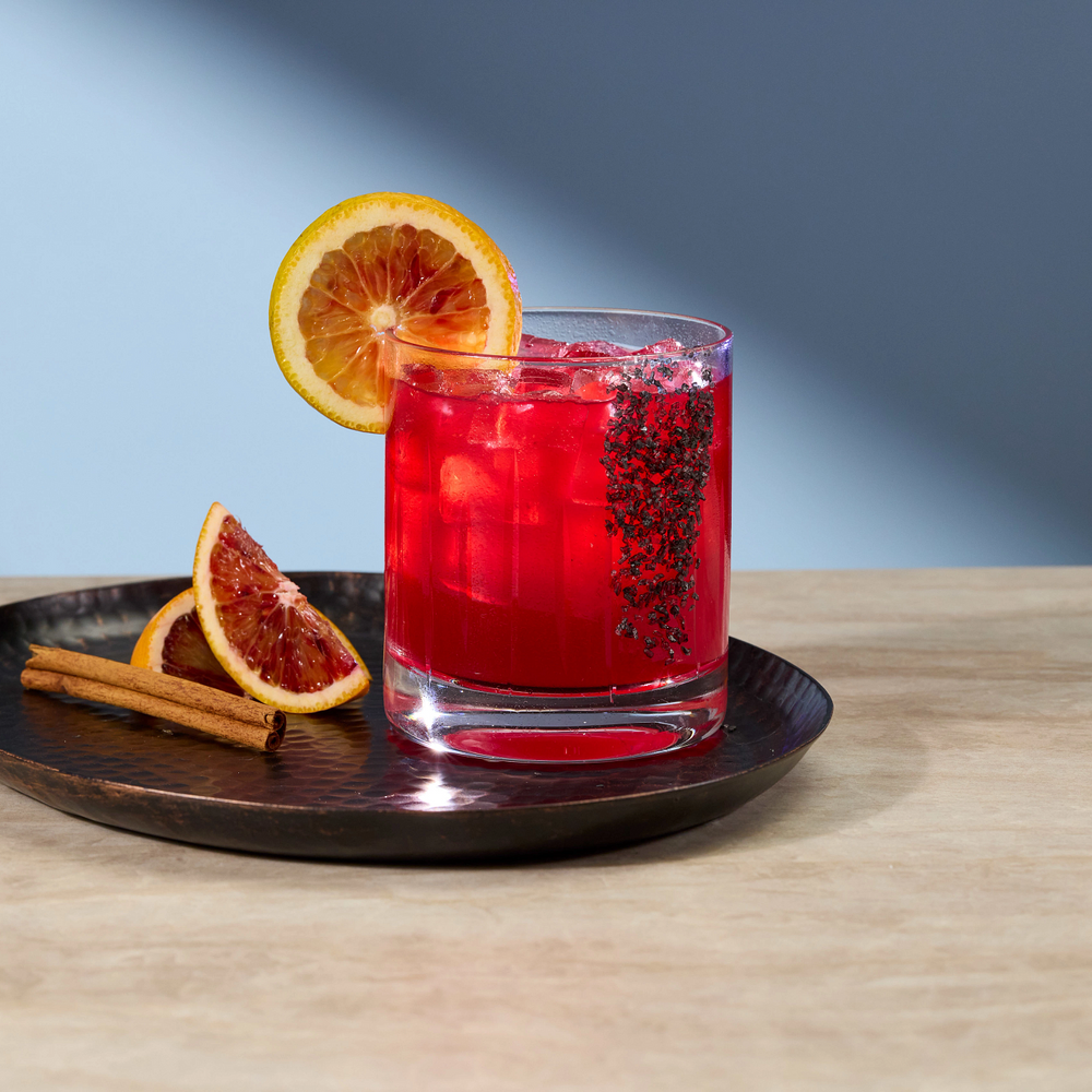 A clear rocks glass filled with a bright red cocktail and ice, garnished with a blood orange slice, set on a dark round tray with two cinnamon sticks and a blood orange slice, on a light wood surface against a blue-gray background.