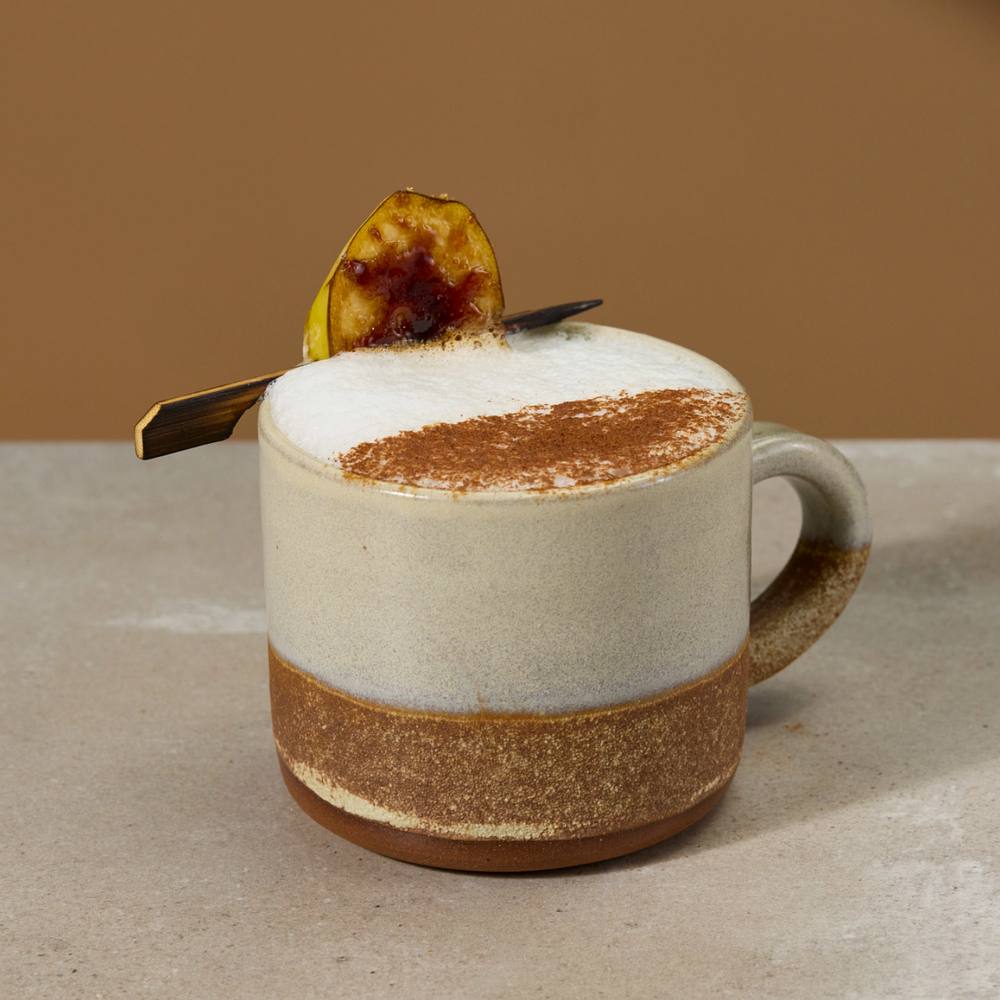 A ceramic coffee mug with a cream-colored upper body, a textured brown band near the base, and a handle on the right side. The mug is filled with a frothy latte topped with cocoa powder and a banana slice garnish, resting on a light stone surface against a warm brown background.