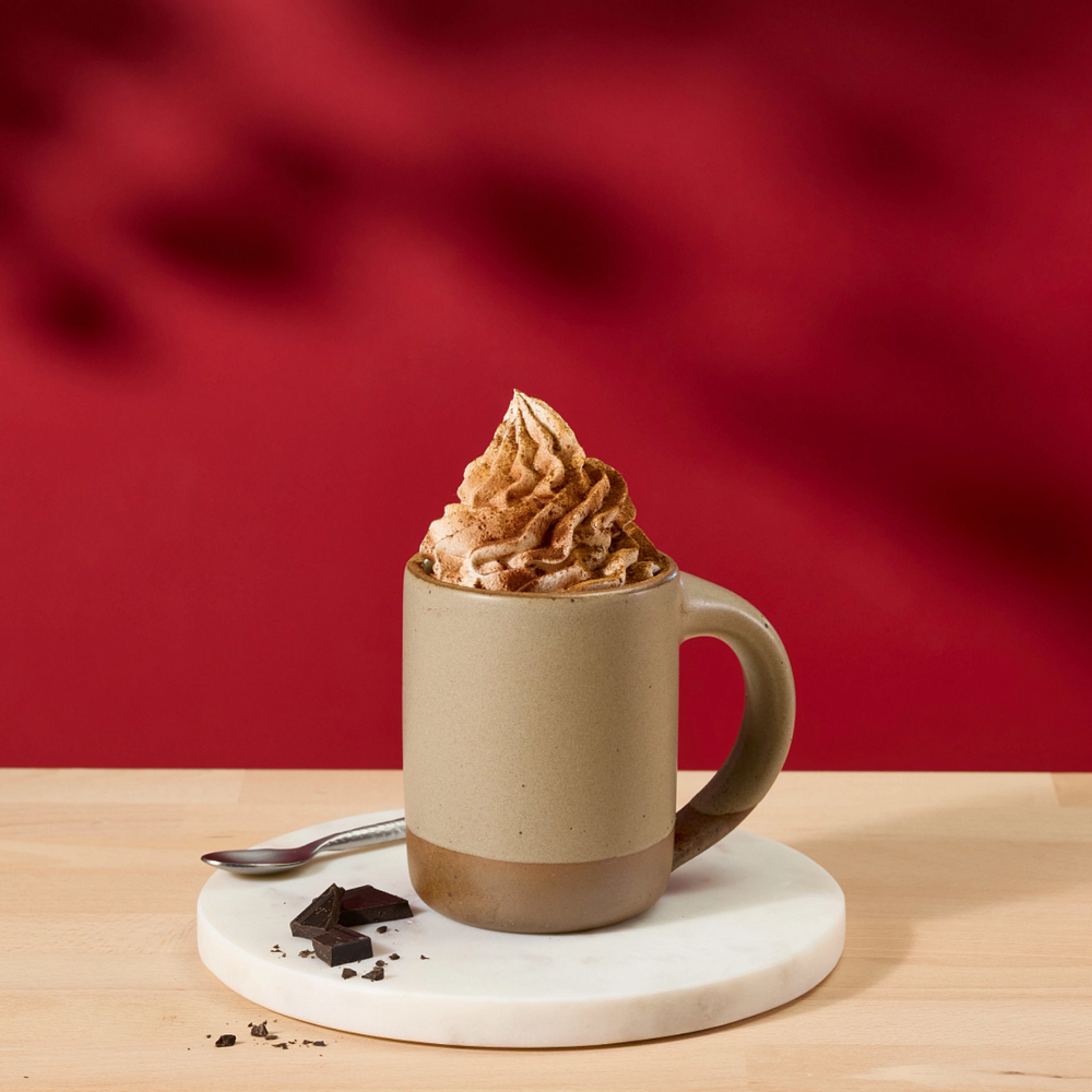 A ceramic mug filled with whipped cream and chocolate drizzle sits on a white saucer with a spoon and chocolate crumbs, set on a light wood table against a deep red background with soft leaf shadows.