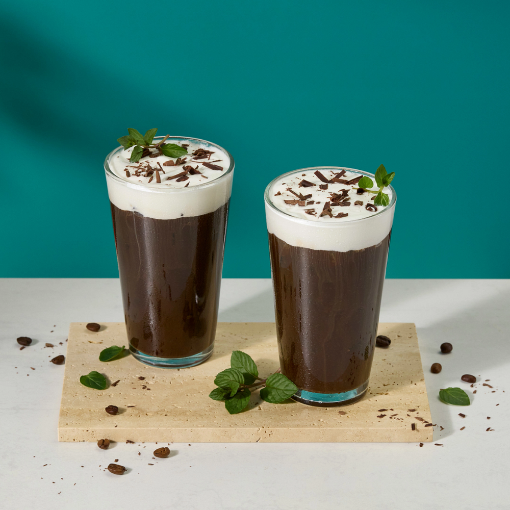 Two tall glasses of chocolate iced coffee with whipped cream and mint garnish, presented on a wooden board with scattered chocolate pieces against a teal background.