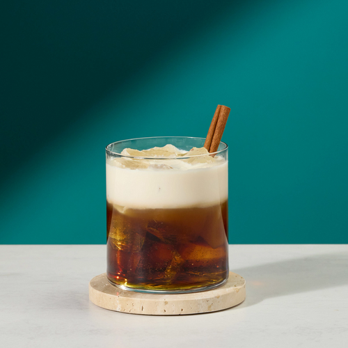 A clear glass iced coffee or tea drink with a thick layer of white foam on top, garnished with a cinnamon stick, sitting on a round coaster on a white surface against a teal background.