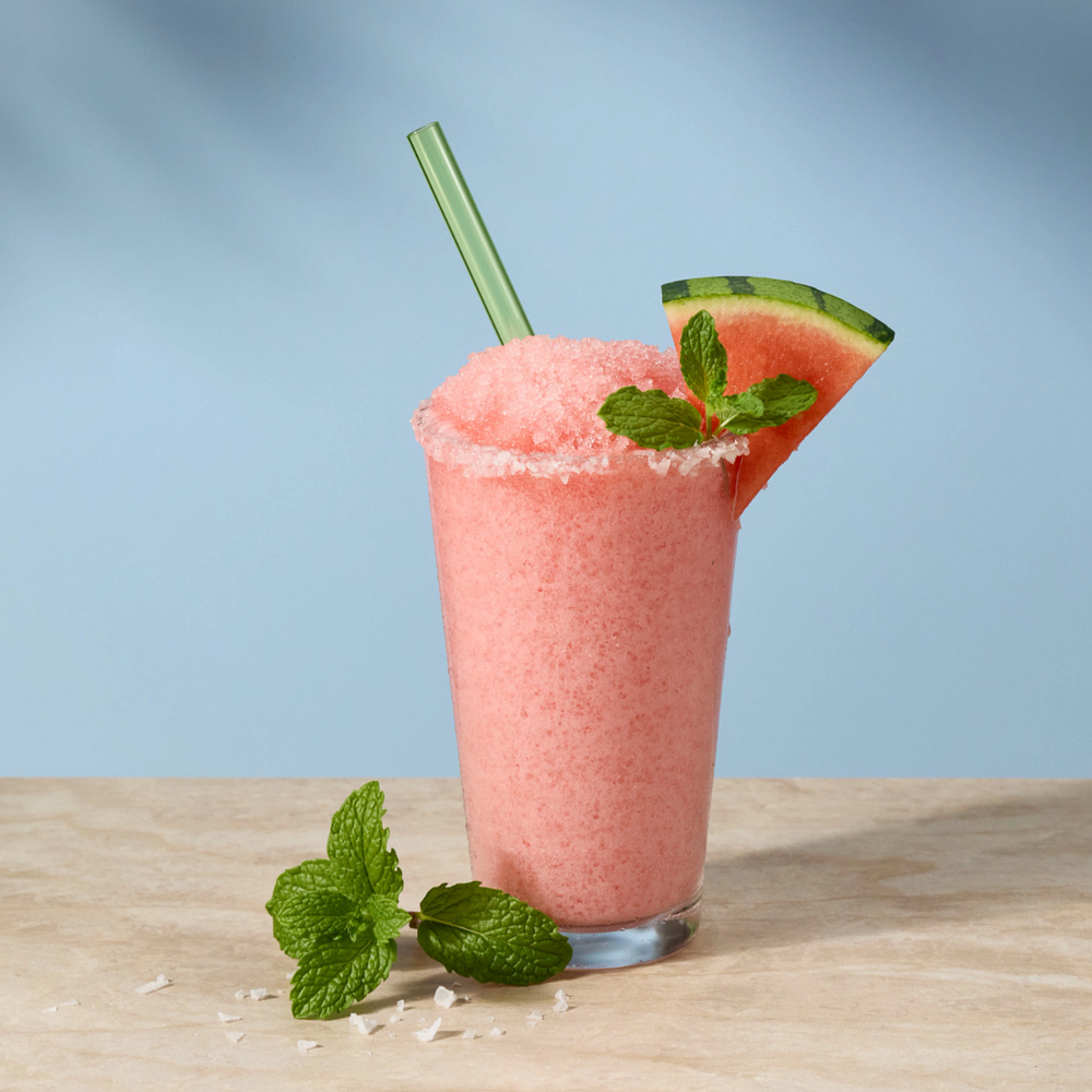 A pink frozen drink in a clear glass with a green straw, garnished with mint leaves and a watermelon slice, set on a light wooden surface against a soft blue background.